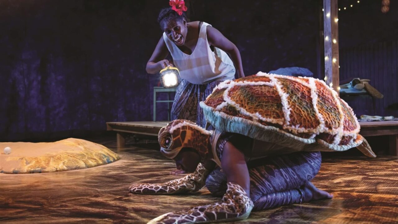 A person in a sea turtle costume is kneeling on a stage floor