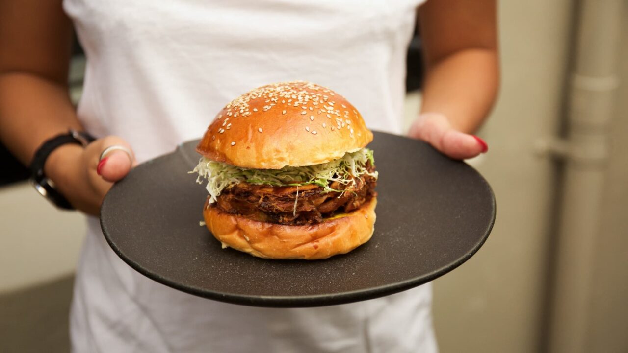 A person presents a sesame-seed bun burger, layered with shredded cabbage and a dark-brown patty, on a dark-grey plate