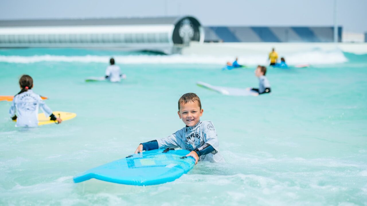 A boy holds a blue surfboard in a wave pool