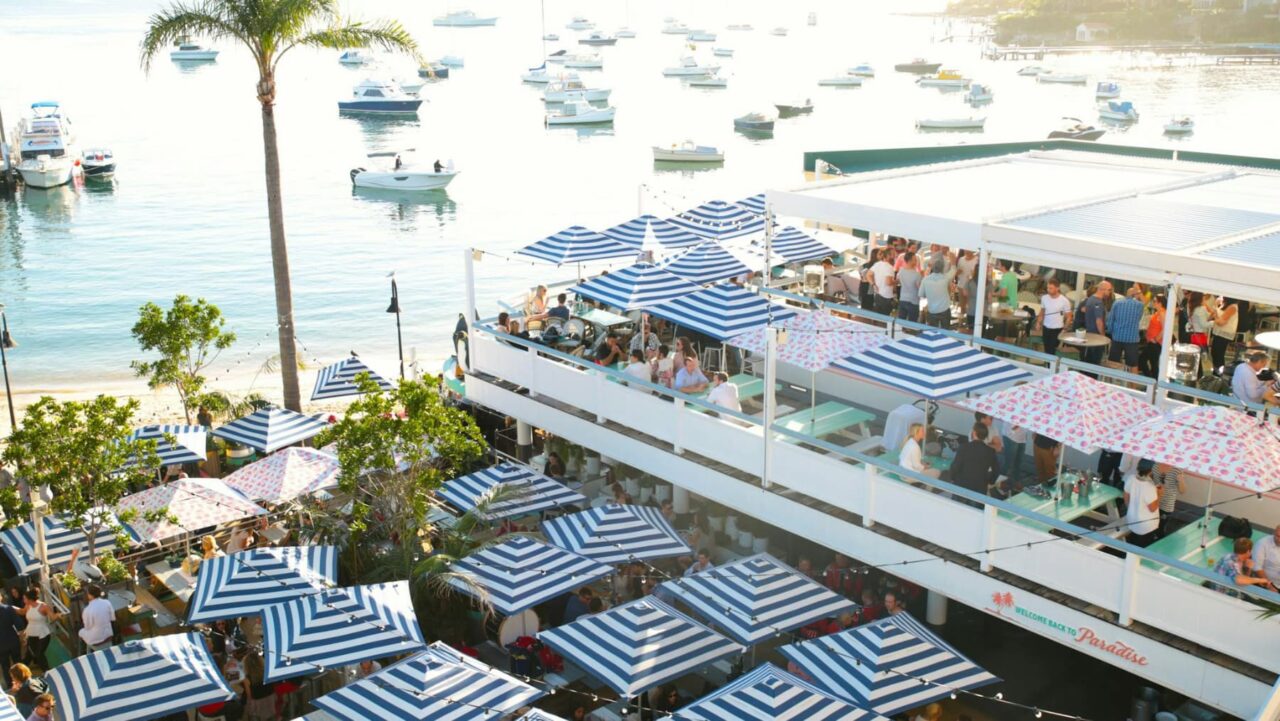 A waterfront restaurant, with many blue and white striped umbrellas, is filled with patrons