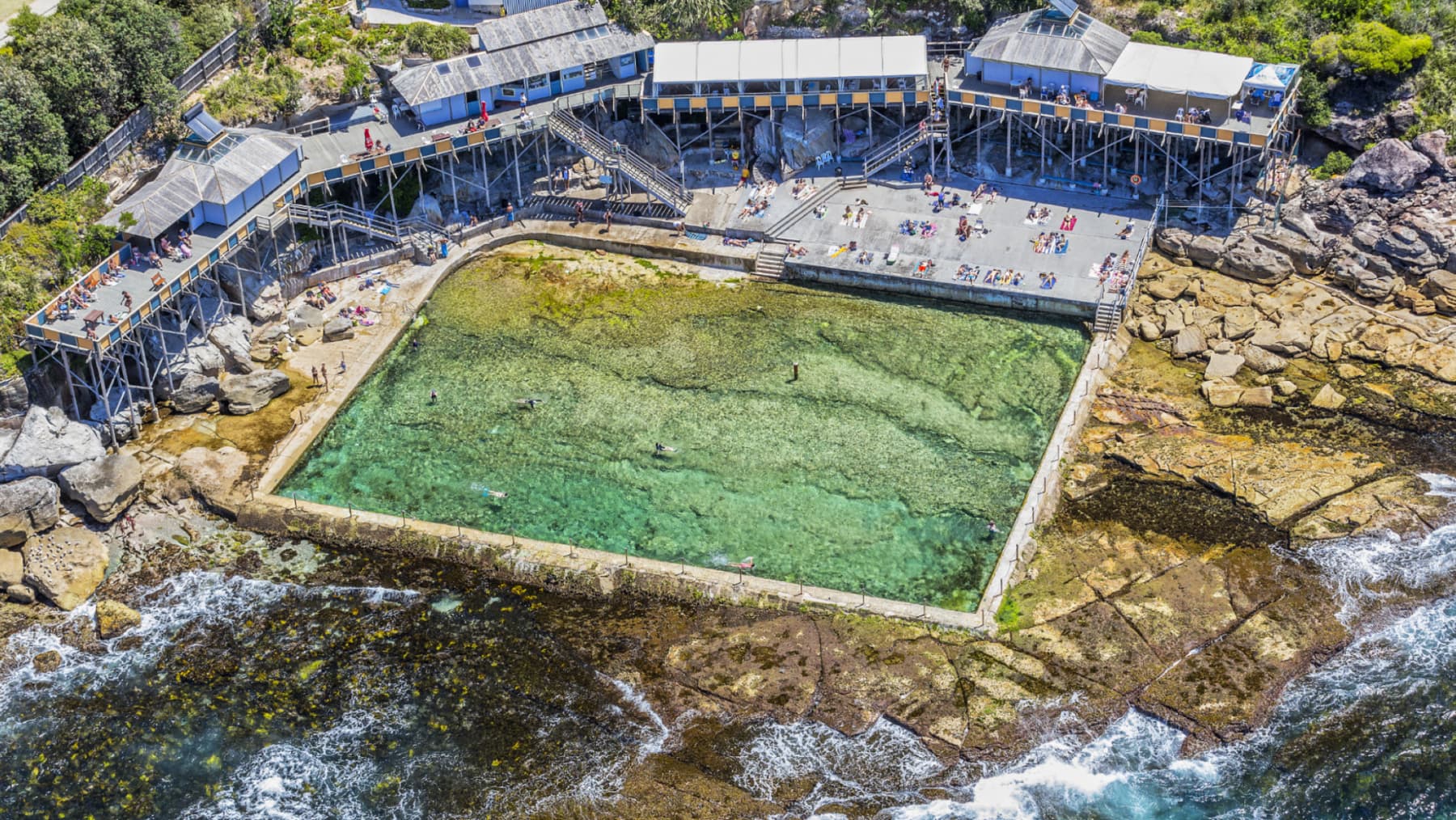 An oceanside pool, filled with clear green water, is surrounded by rocky terrain and wooden structures