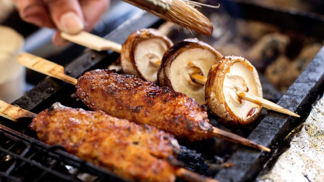 Meat and mushroom skewers are being brushed with sauce while grilling on a hot grate