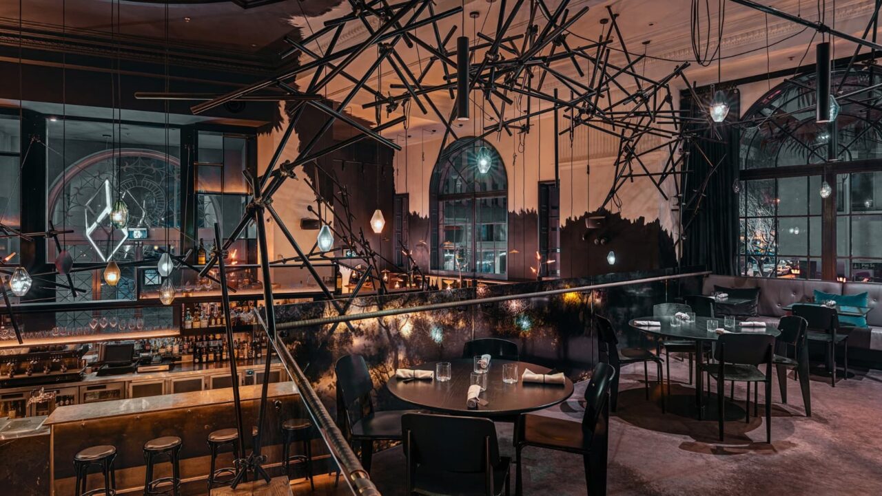 A complex light fixture hangs above dining tables and a bar, within a dimly lit, upscale restaurant setting
