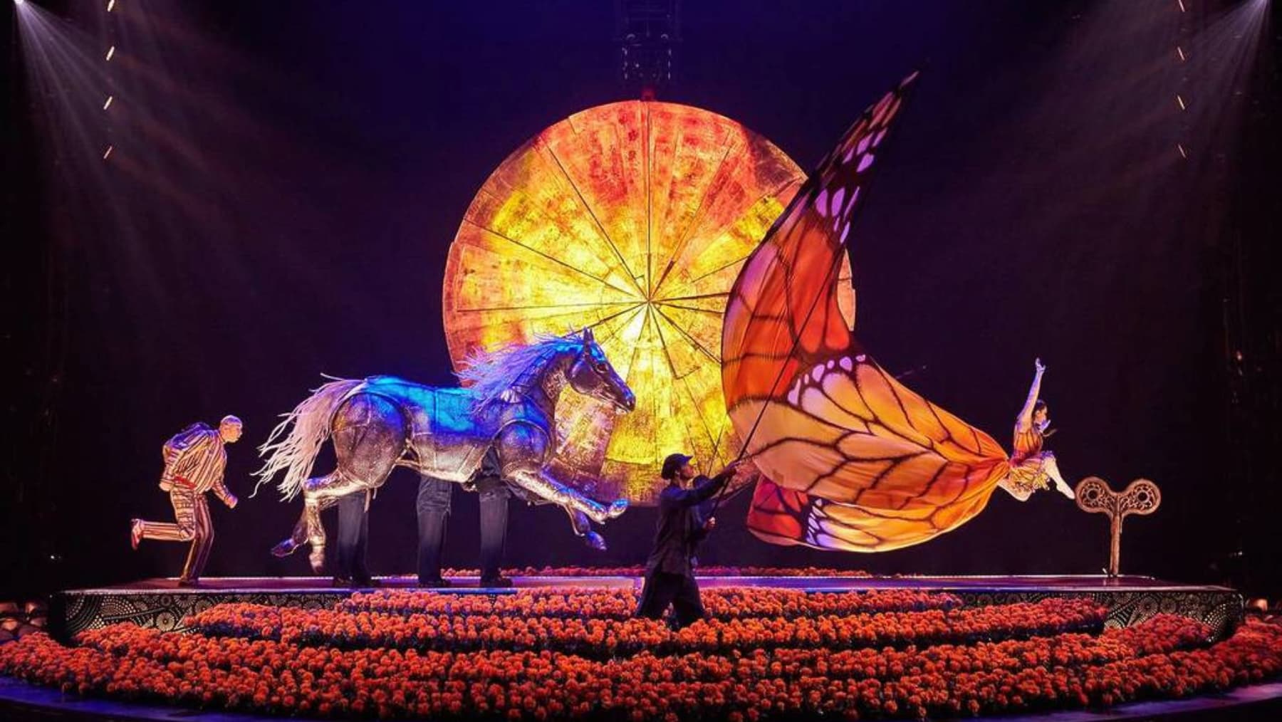 A mechanical horse and a large butterfly puppet are manipulated by performers on a stage covered in orange flowers, set against a large illuminated circular backdrop