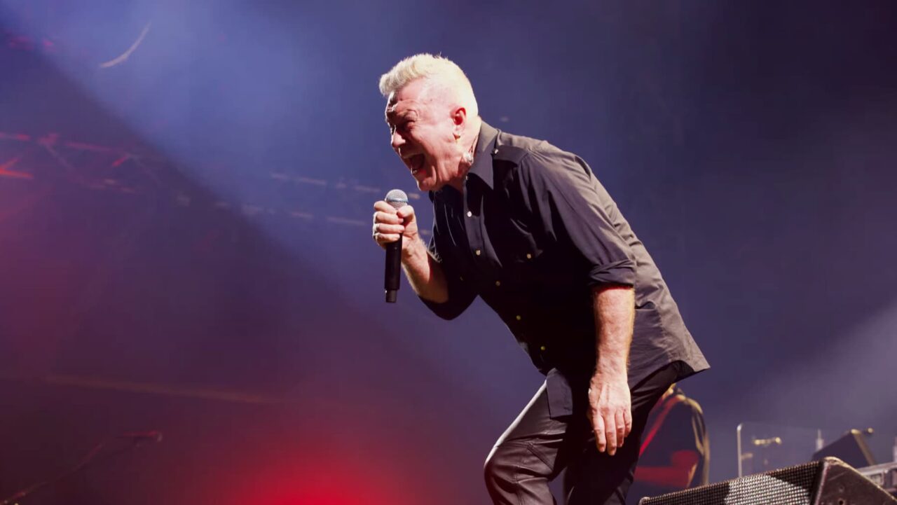 A man, singing intensely into a microphone, performs on a dimly lit stage