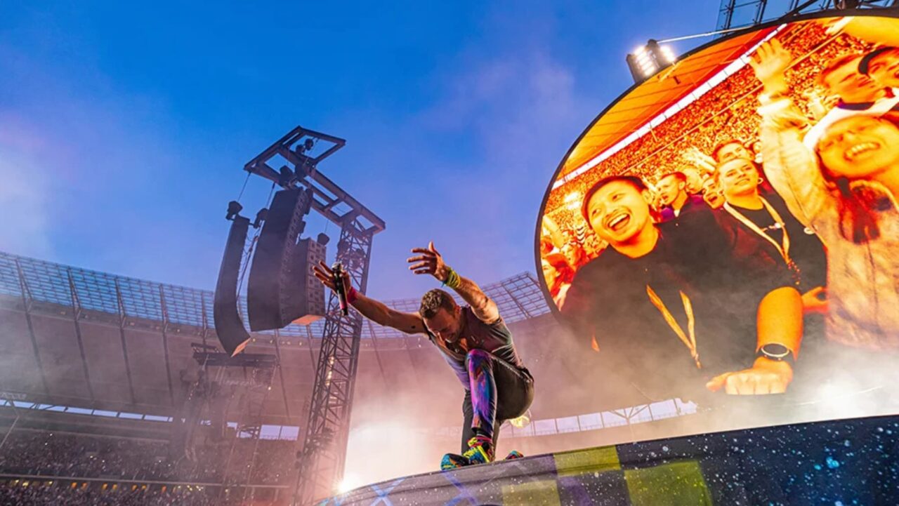 A male performer, dynamically posing with a microphone, is center stage at a stadium concert
