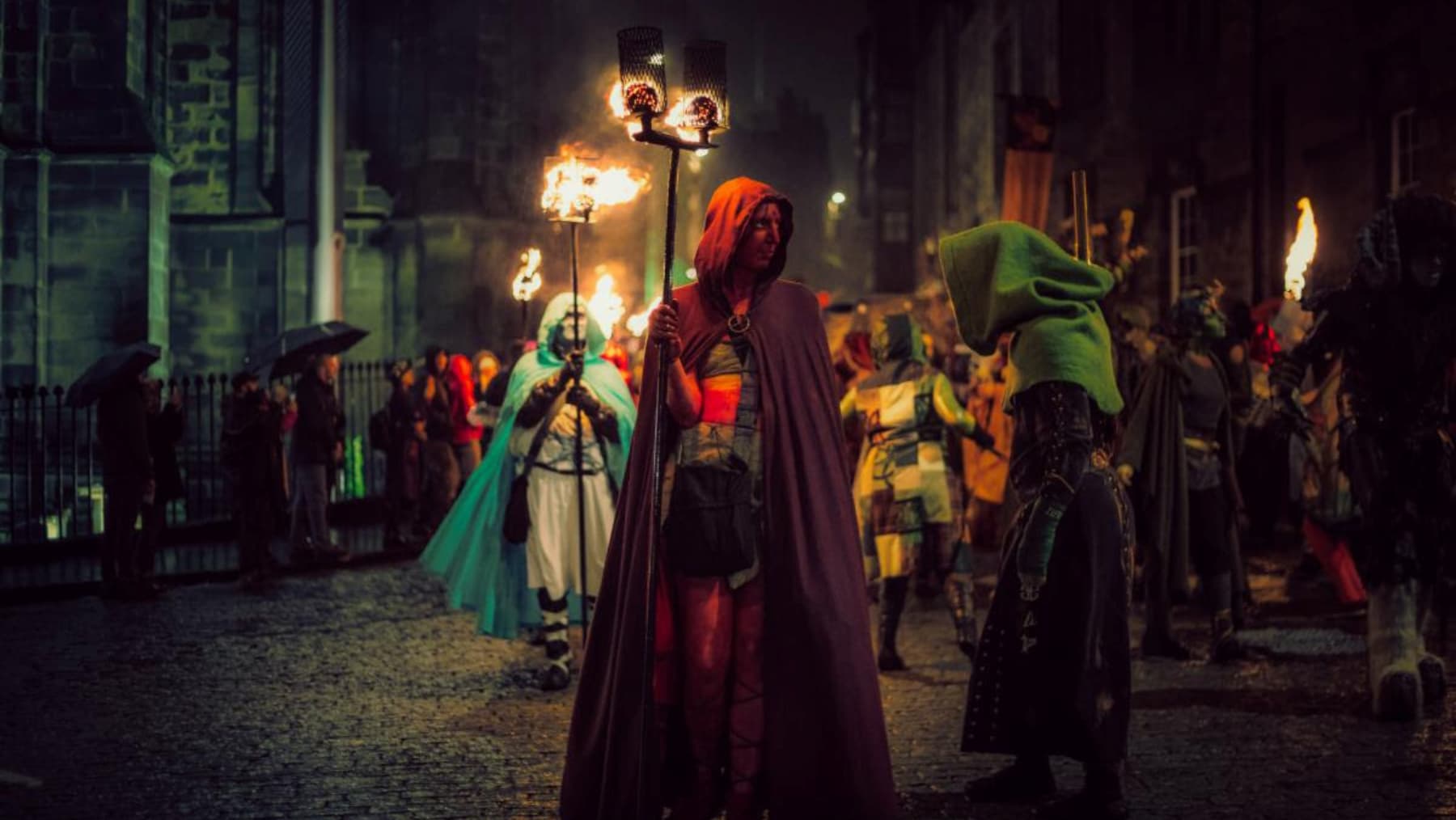 Costumed figures, carrying flaming torches, process through a dark, cobblestone street at night