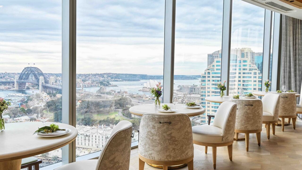 Elegant tables and chairs sit near expansive windows overlooking Sydney's harbor, bridge, and Opera House