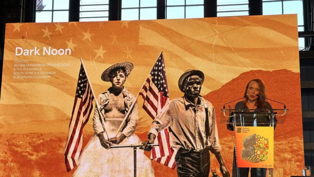 A woman speaks at a podium, behind which a large poster depicts three figures carrying American flags in a desert landscape