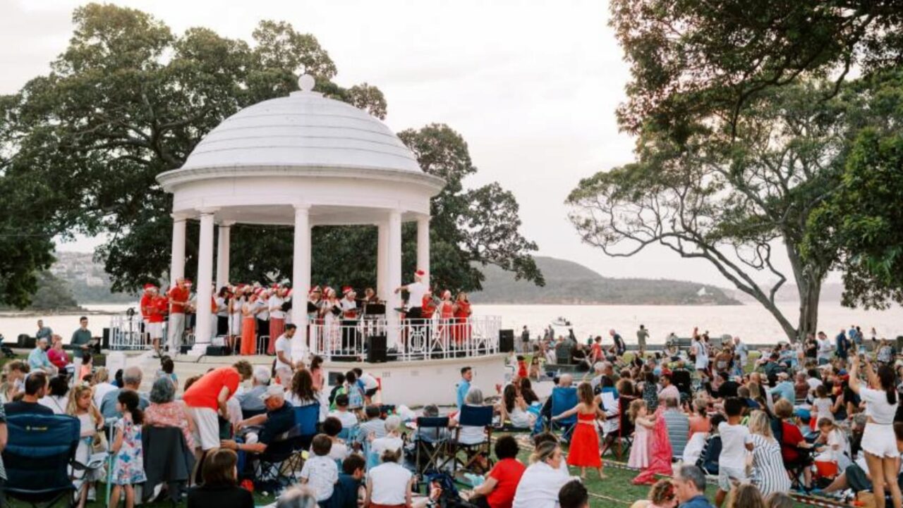 A choir performs on a gazebo stage, in a park setting, near a body of water, for a large audience seated on the grass