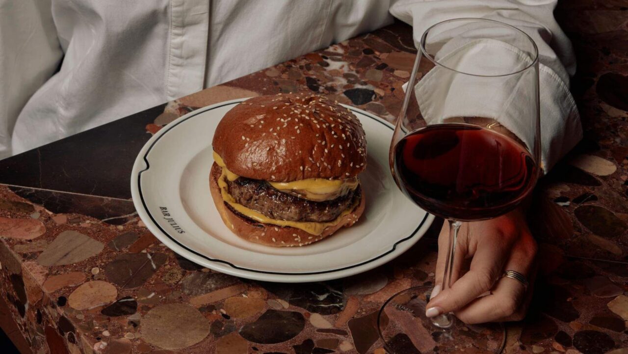 A cheeseburger rests on a plate marked "BAR JULIUS," accompanied by a glass of red wine, all situated on a terrazzo table