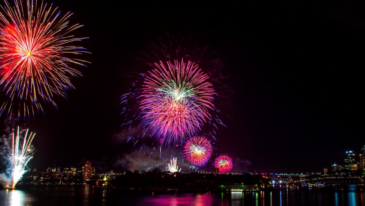 Multicolored fireworks explode over a cityscape at night, reflecting in dark water