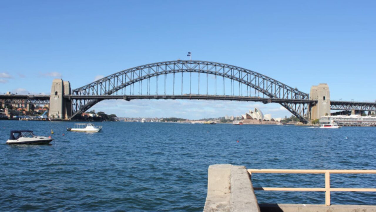 A steel arch bridge spans a body of water, with boats floating below