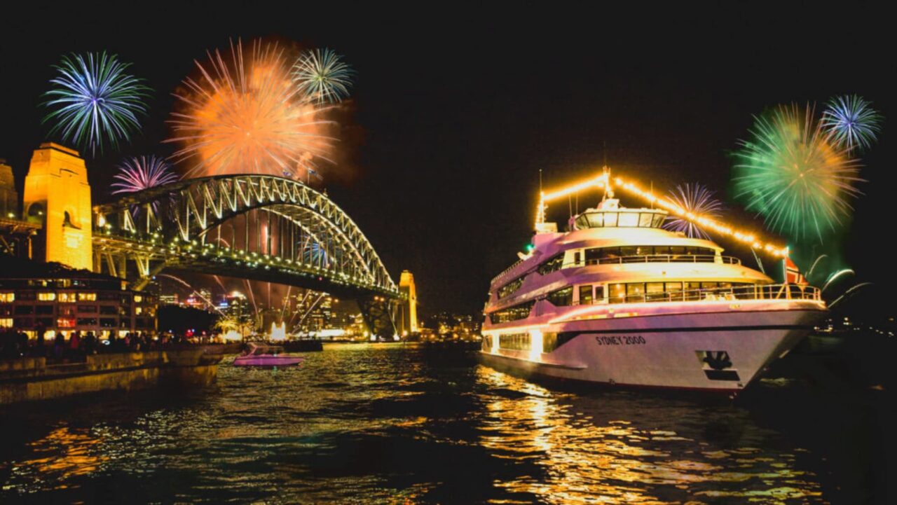 A brightly lit yacht sails under a bridge during a fireworks display