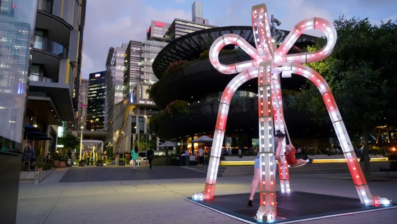 A large illuminated bow-shaped swing is being used by a woman and child; the setting is an urban plaza at dusk, with modern buildings and city lights in the background