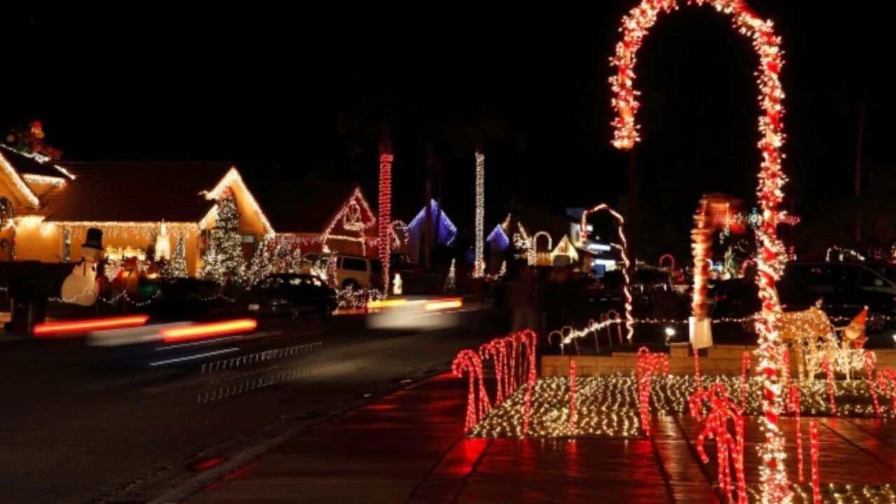 A large illuminated candy cane stands on a residential street, decorated with Christmas lights
