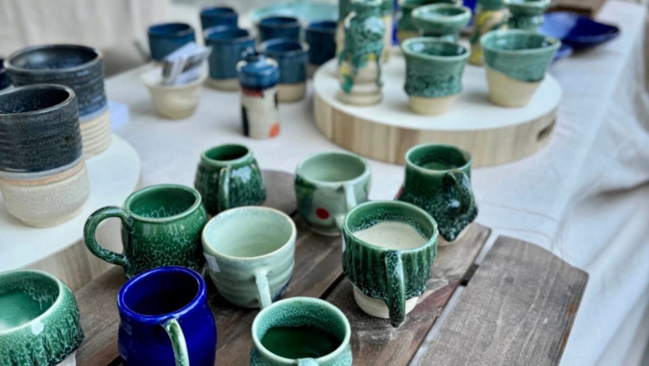Many handcrafted mugs and bowls sit on wooden surfaces, displayed at an outdoor market
