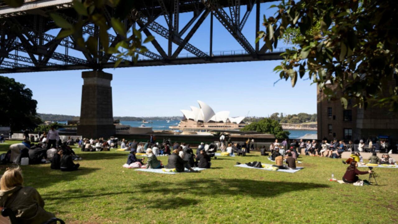 People relax on blankets, picnicking on a grassy area under a bridge