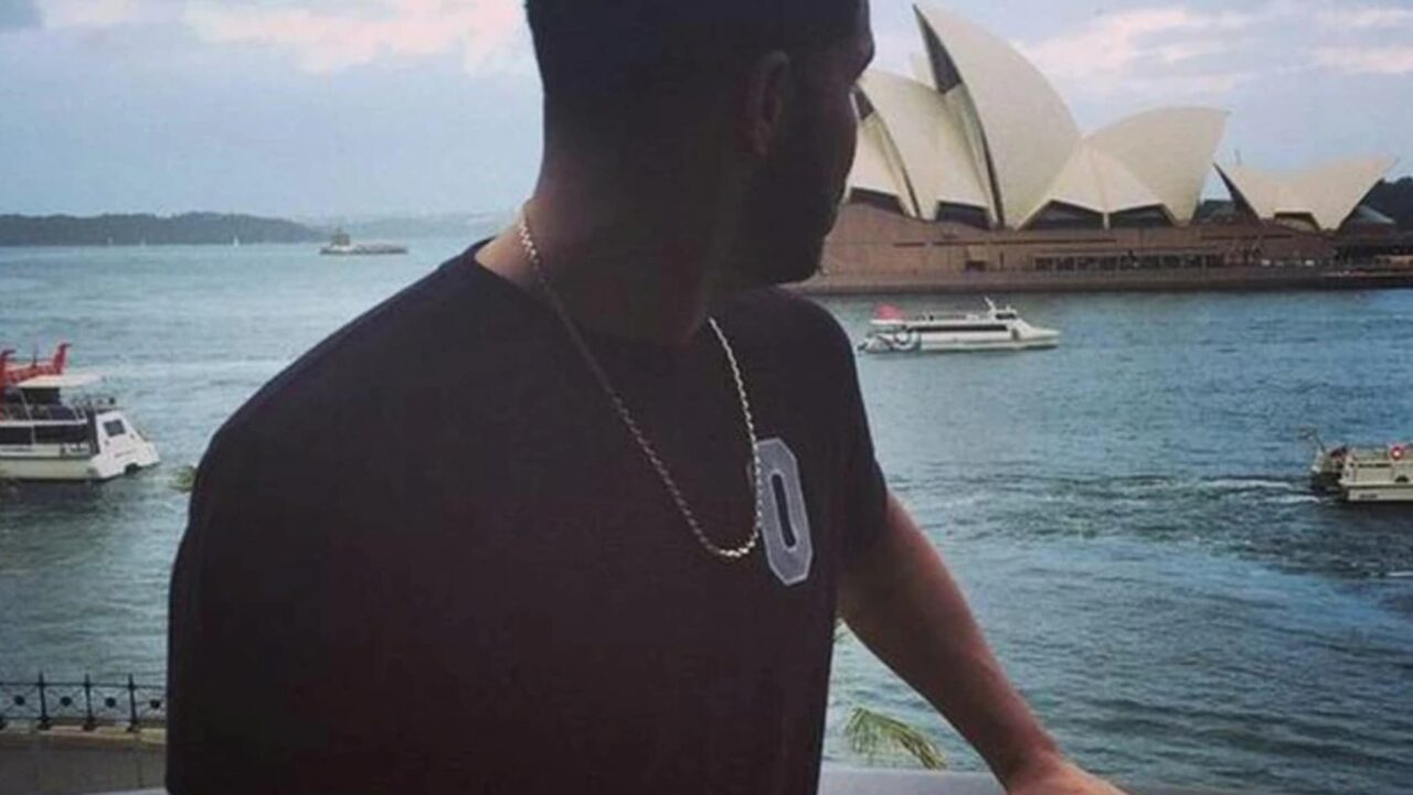 A man, wearing a black shirt and a necklace, stands overlooking a harbor; the Sydney Opera House is visible in the background