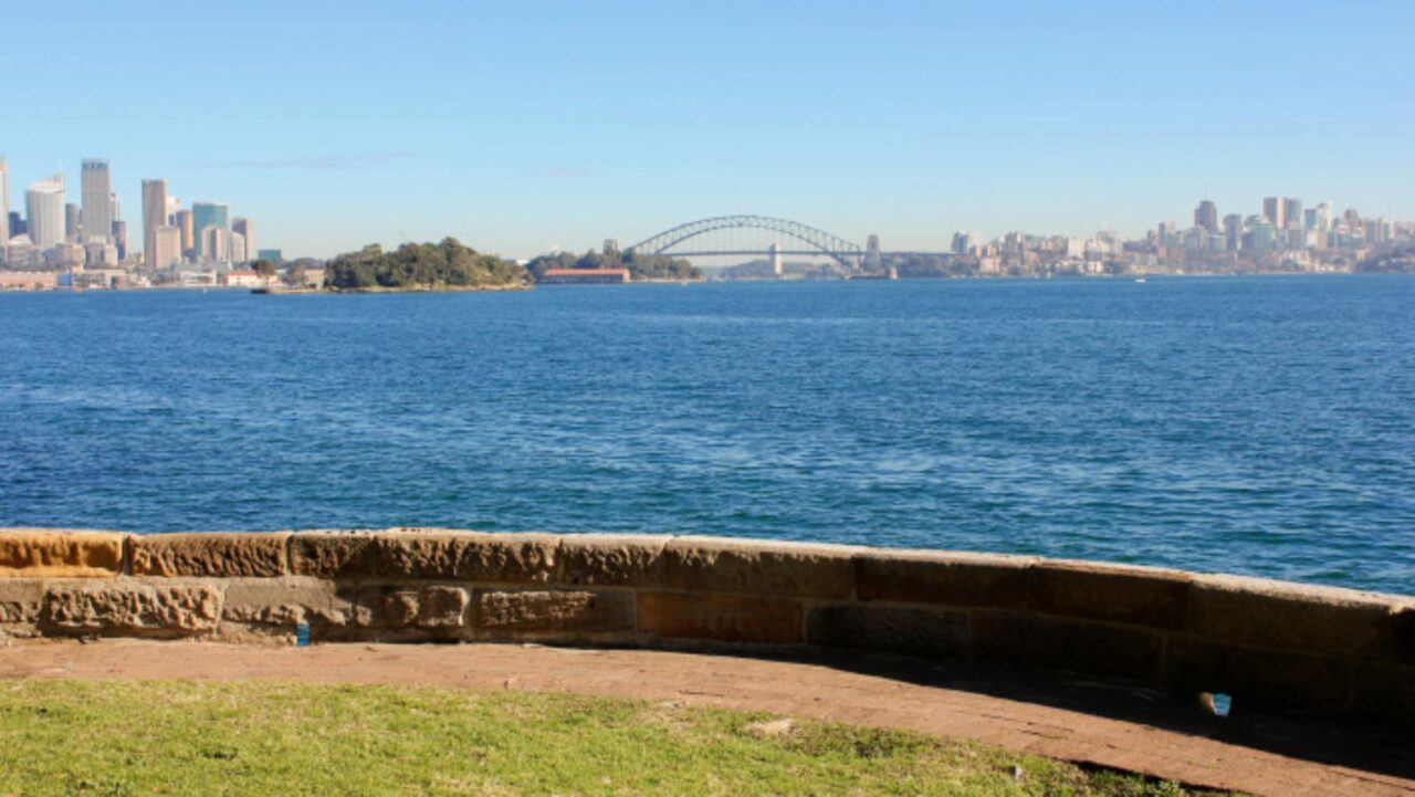 A low stone wall overlooks a calm body of water; a city skyline and bridge are visible in the distance under a clear sky