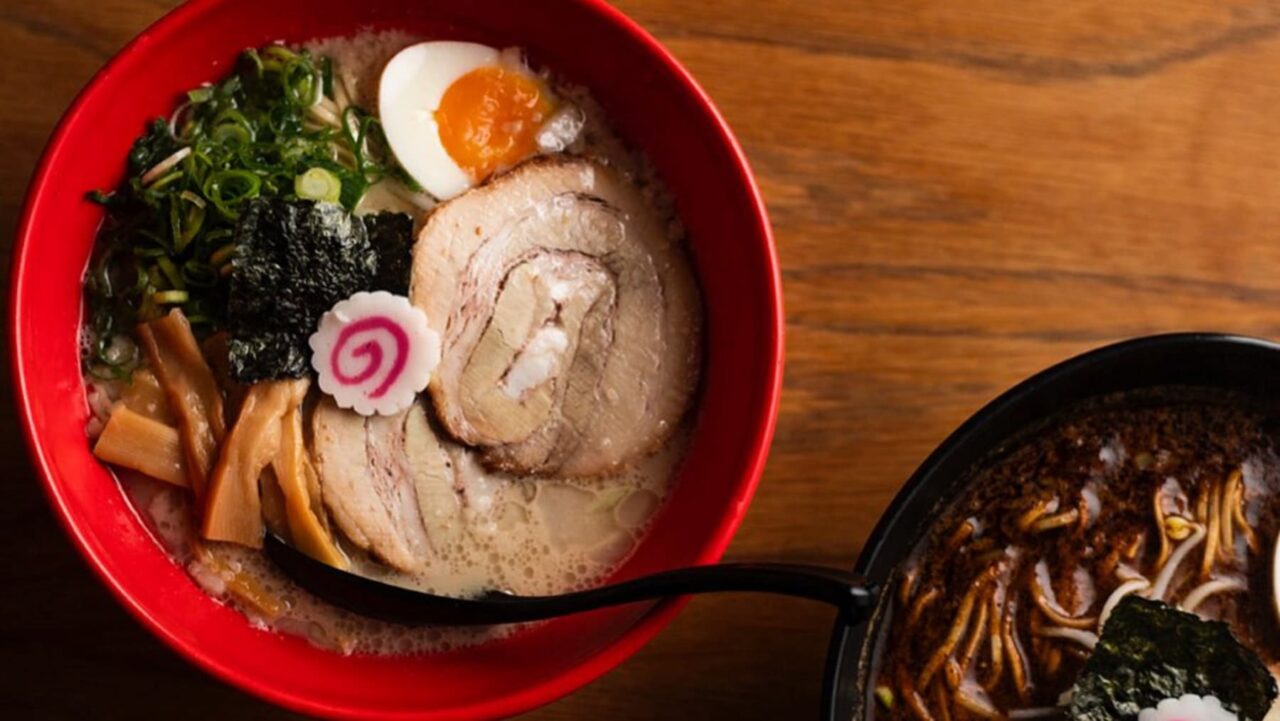 A bowl of ramen, containing pork slices, a soft boiled egg, nori, and other toppings, sits on a wooden table