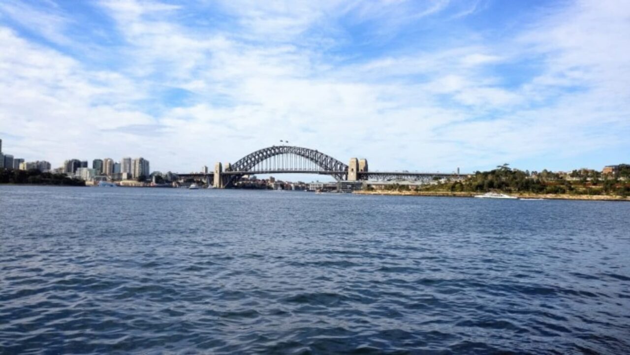 A steel arch bridge spans a body of water, connecting a city skyline under a partly cloudy sky