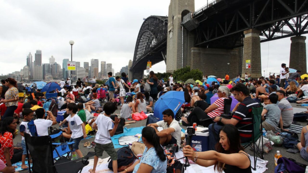 A large crowd sits on blankets and chairs, under umbrellas and small tents