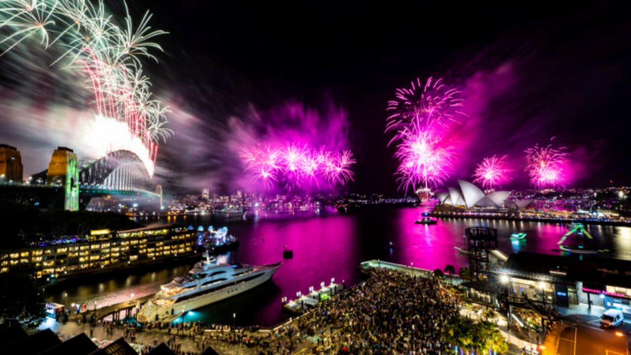 Fireworks explode over Sydney Harbour Bridge and Opera House, illuminating a large crowd gathered below on the waterfront