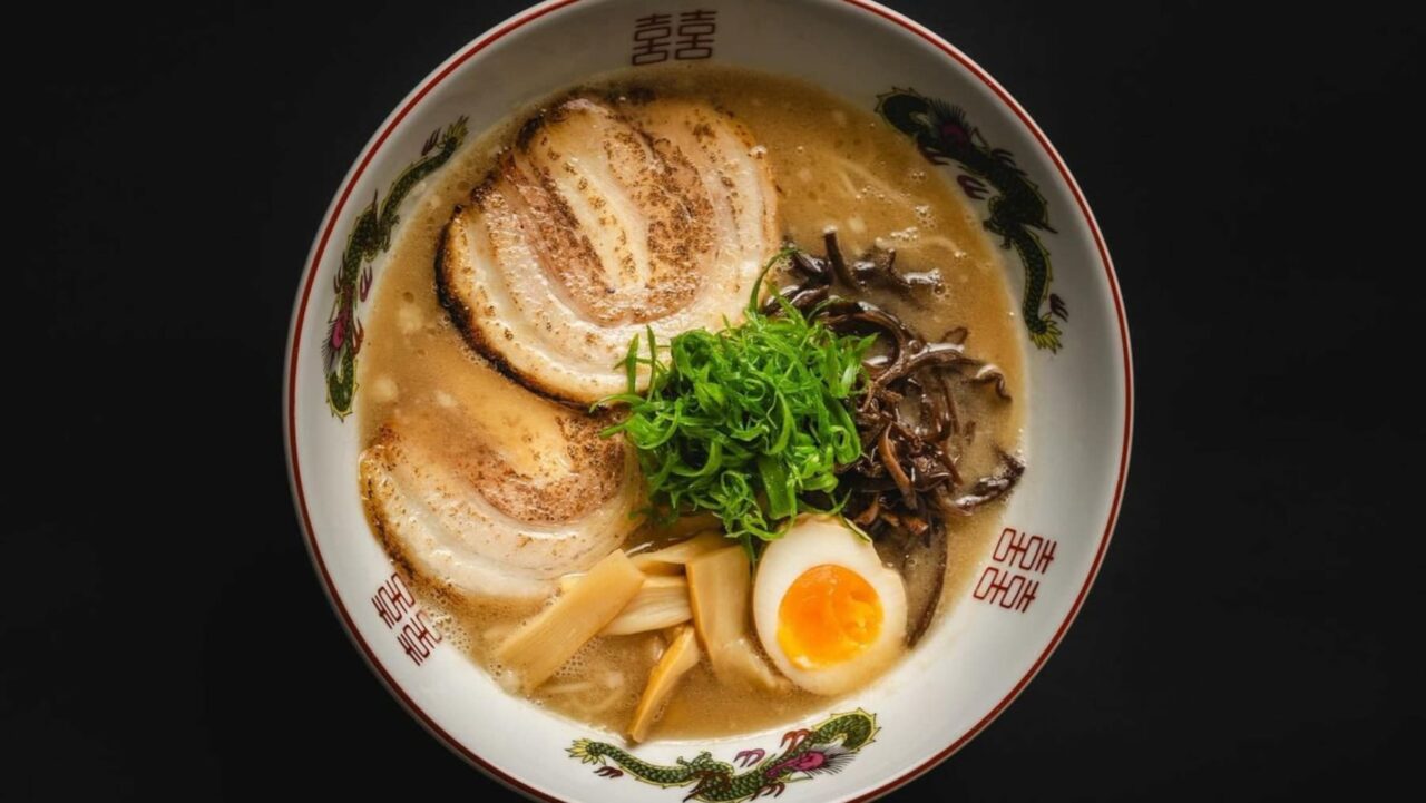 A bowl of ramen, topped with chashu pork, a soft boiled egg, bamboo shoots, wood ear mushrooms, and scallions, sits on a dark surface