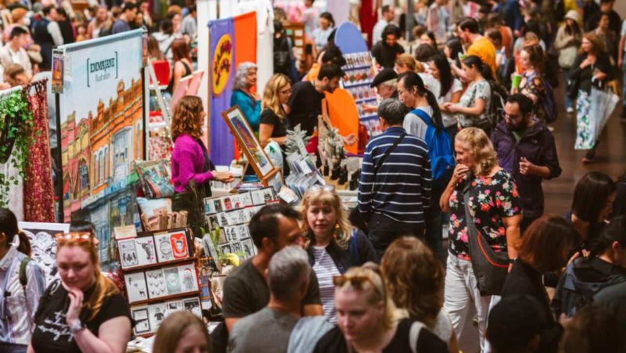 A craft market displays various goods; many people browse and shop amidst colorful banners and stalls