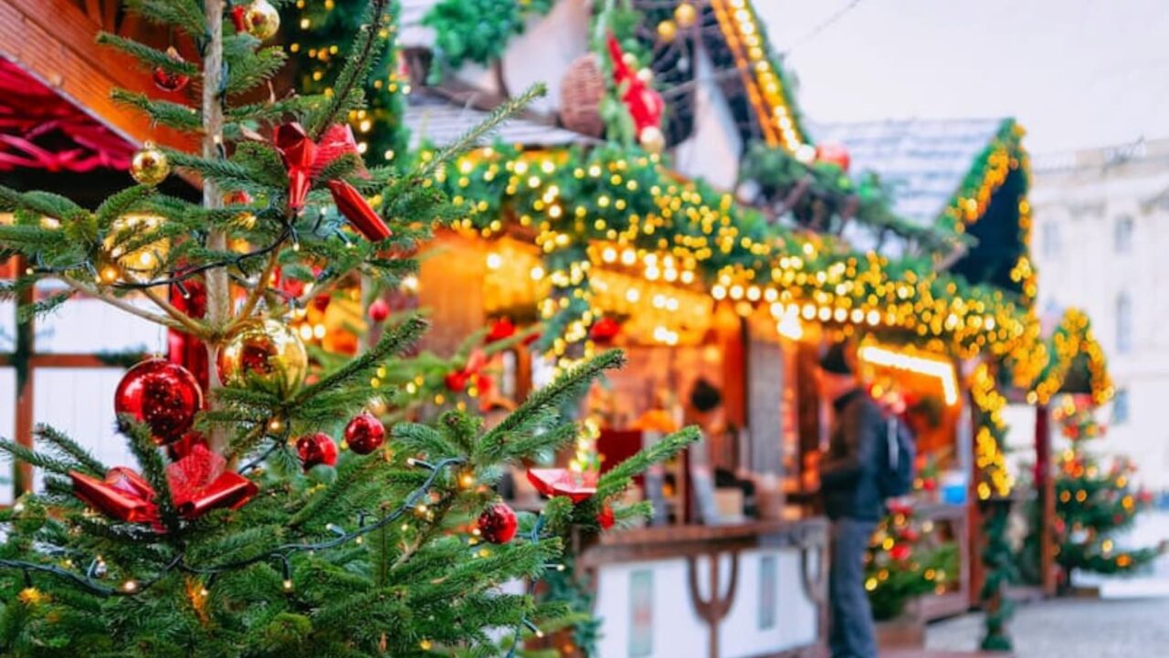 A decorated Christmas tree, adorned with red ornaments and lights, stands before festively lit market stalls