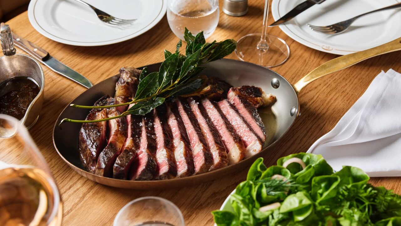 A sliced steak rests in a copper pan, garnished with bay leaves
