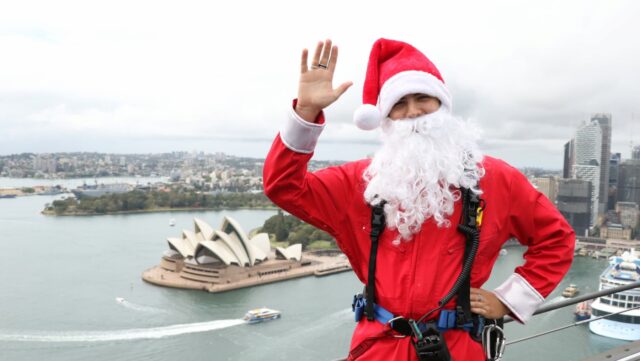 A man dressed as Santa stands on top of the Sydney Harbour Bridge with Sydney Opera House in the background- things to do in winter in sydney