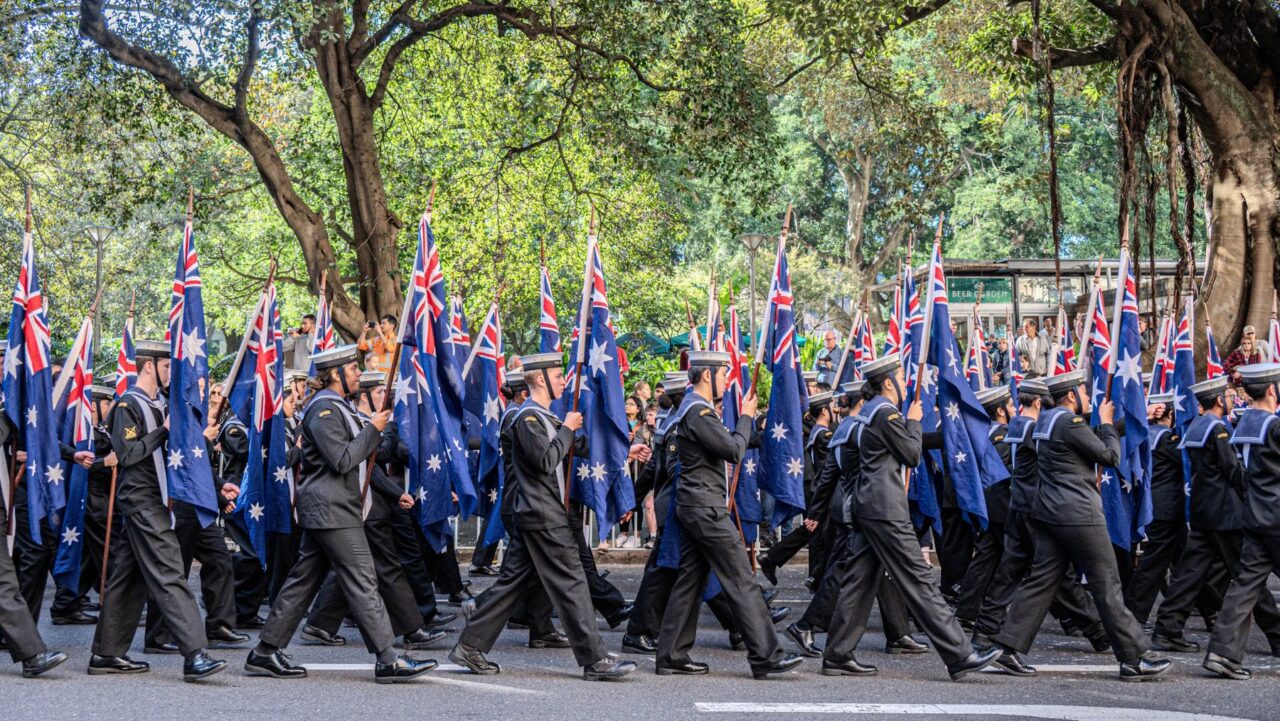 A large group of service men and women in uniform march carrying Australian flags