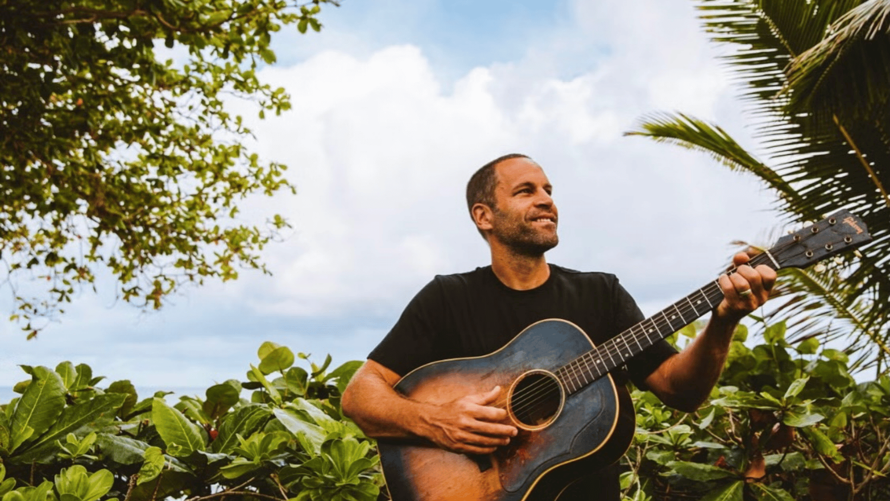Musician Jack Johson holds a guitar in a tropical setting