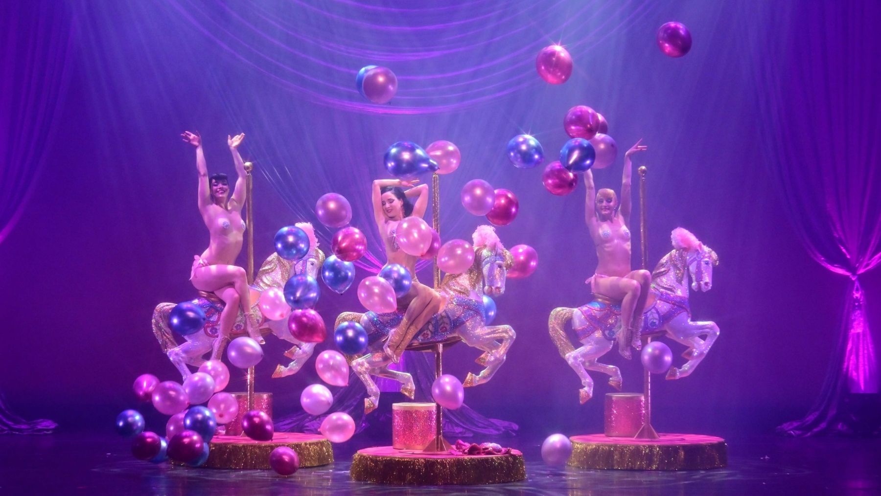 3 burlesque dancers on stage with carousel horses and balloons