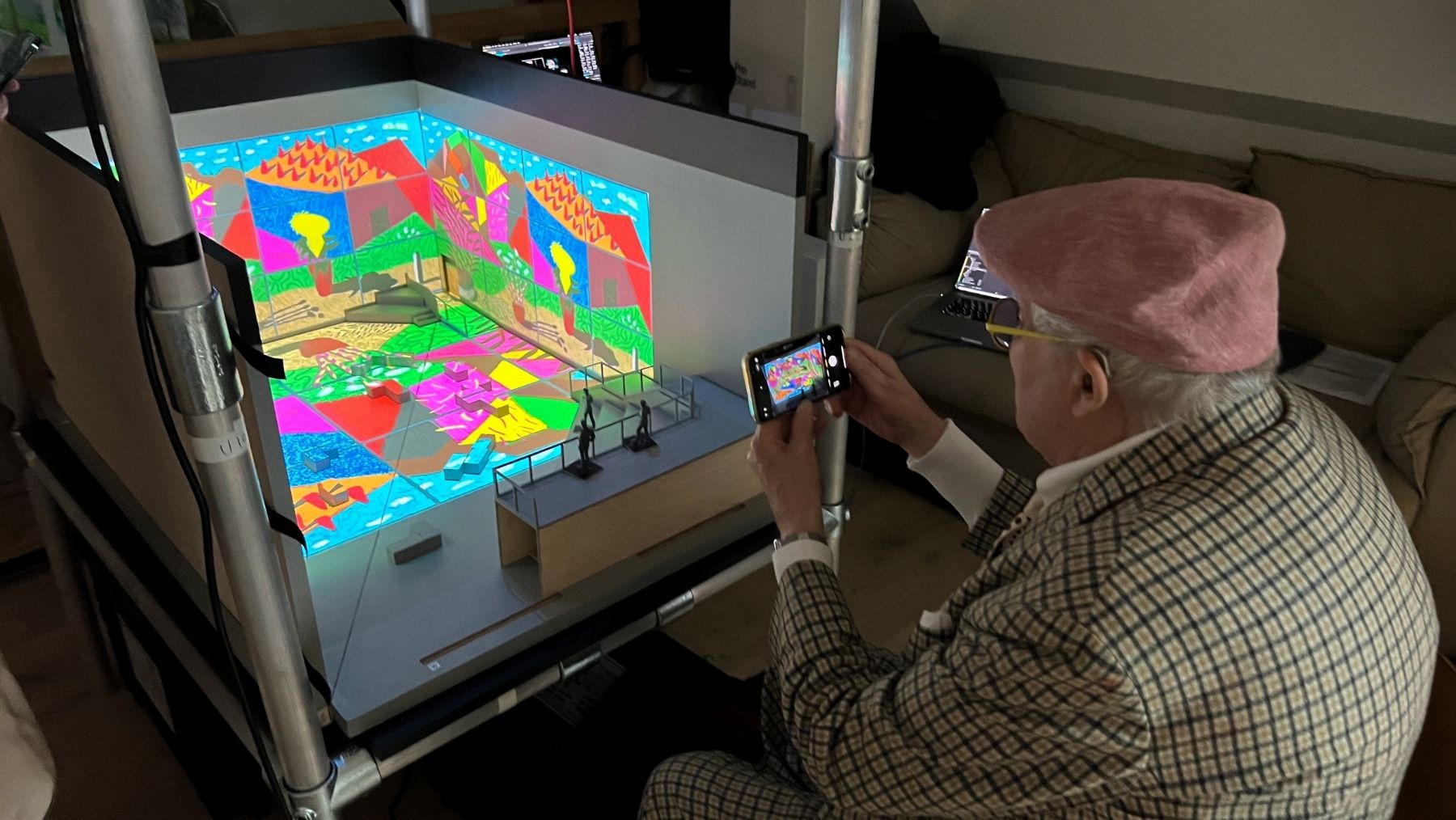 Artist David Hockney in a cap looks at an image on his phone that is capturing a model of his exhibition in front of him.