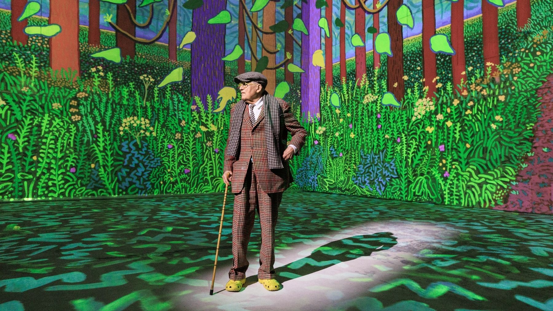 A man stands in a check suit holding a cane and wearing crocs in front of a colourful art projection